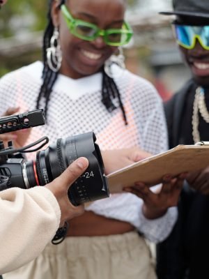 Filming crew holding camera and reviewing script on clipboard during an outdoor film shoot. Team members focused on scene planning without direct eye contact with camera